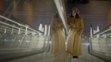 Modern bir metro istasyonunda şık bej bir trençkot ve şapka giyen bir kadın dinamik bir yürüyüş yolunda yürüyor.