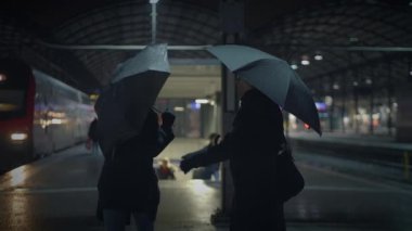 Bir çift, gece loş bir tren istasyonunda romantik ve atmosferik bir sahne oluşturan bir şemsiyeyi paylaşıyor..