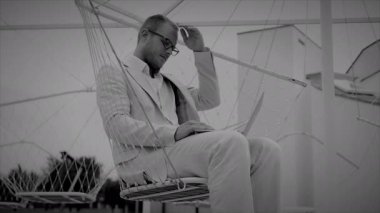 Beyaz takım elbiseli, siyah beyaz gözlüklü bir adamın düşünceli bir şekilde salıncakta oturuşu..