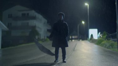 Yağmurda şemsiyeyle sokak lambalarının altında duran figür gizemli bir şehir atmosferi yaratıyor.
