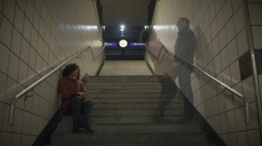 Gecenin karanlığında loş bir metro terminalinde, yalnız bir kadın merdivenlerde oturur, şehir yalnızlığını ve izolasyonu simgeler.