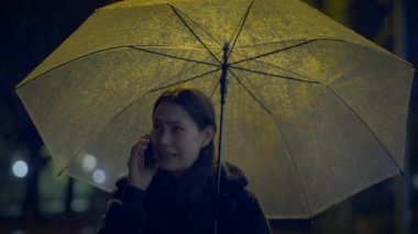 Sarı sokak lambalarıyla dolu yağmurlu bir gecede şeffaf şemsiye altında telefonla konuşan kişi.