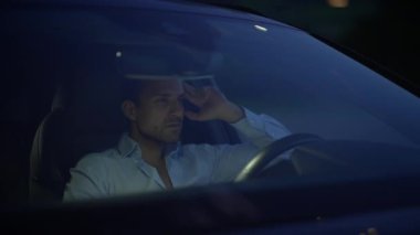 Bir adam gece arabasında oturuyor, derin düşüncelere dalmış, ortam ışıklarının yumuşak parıltısıyla çevrili.