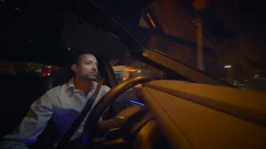 Bir adam gece parıldayan şehirde araba sürüyor, parlak ışıkları ve yansıtıcı iç mekanı aydınlatıyor.