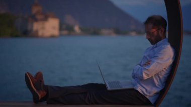 Bir adam gün batımında sakin bir göl kenarında huzur içinde otururken dikkatle dizüstü bilgisayarına odaklanır.
