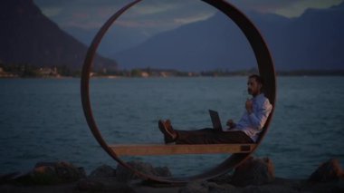 Bir insan suyun yanındaki dairesel metal bir yapıda otururken bir dizüstü bilgisayar üzerinde huzur içinde çalışıyor.