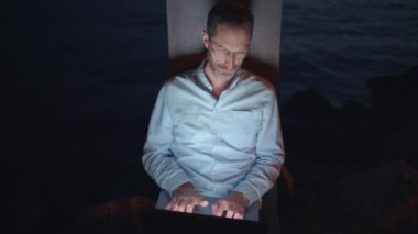 Odaklanmış bir adam, modern dijital göçebe yaşamını somutlaştıran, sakin deniz kenarındaki dizüstü bilgisayarında çalışır.
