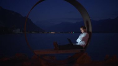 Huzurlu bir gece sahnesi. Dairesel bir kurulum içinde huzurlu bir göl kenarında dizüstü bilgisayar üzerinde çalışan bir kişi.