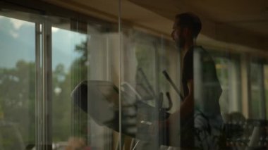 Koşu bandında egzersiz yapan odaklanmış bir birey, büyük pencereler dışarıda güzel bir doğa sergiliyor.