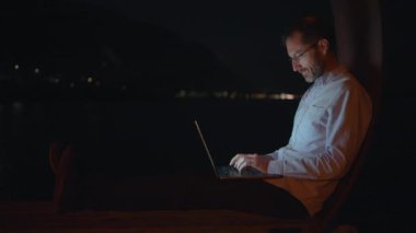 Gece geç saatlere kadar laptopuyla çalışan bir adam huzurlu ve güzel bir rıhtımın yanında rahatça oturuyor.