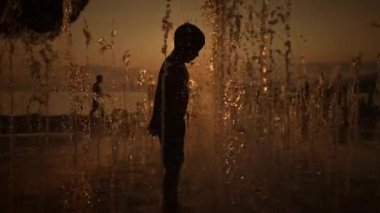 Gün batımında fıskiyeden gelen ferahlatıcı spreyin keyfini çıkaran bir çocuğun hoş bir silueti.