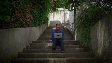Yeşilliğin ortasında beton merdivenlerde rahatça otururken dikkatle dizüstü bilgisayarına odaklanmış bir birey.
