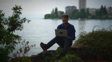 Bir adam deniz kenarında huzur içinde oturur, dizüstü bilgisayarına odaklanır ve etrafını dingin bir ortam sarar.