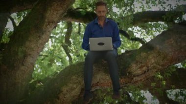 Bir adam bilgisayarını kullanarak sağlam bir ağaç dalında rahatça otururken uzaktan kumandalı bir işle meşgul olur.