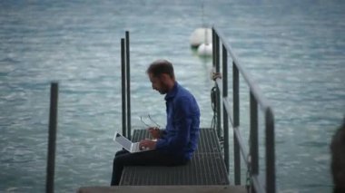Bir adam sakin sulara tepeden bakan bir rıhtımda rahatça otururken dizüstü bilgisayarına derinden odaklanır.