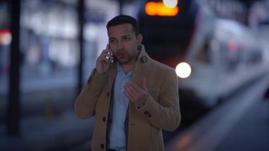 Akşamları tren istasyonunda bir telefon görüşmesi yapan bir adam. Arka planda bir tren var..