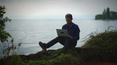 Bir adam, sakin göl kenarında dizüstü bilgisayarı üzerinde özenle çalışıyor ve doğanın ortasındaki uzak işlerin güzelliğini gösteriyor.
