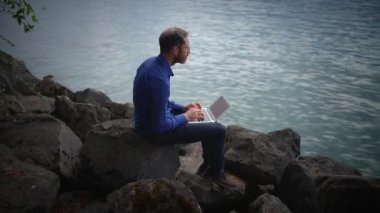 Sakin sularla çevrili kayalık bir sahilde dizüstü bilgisayarla oturan bir insan, uzaktan çalışmayı ve üretkenliği simgeliyor.