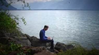 Odaklanmış bir birey, sakin ve sakin bir gölün sakin sularında otururken dizüstü bilgisayar kullanıyor.