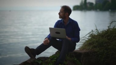Kendini işine adamış ve işine odaklanmış bir adam göl kenarında dizüstü bilgisayar kullanıyor.