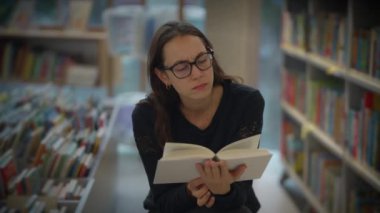 Odaklanmış bir kadın, yüksek raflarla dolu huzurlu bir kütüphanede kitap okumaya dalmış.
