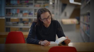 Sayısız kitapla çevrili modern bir kütüphanede özenle ve dikkatle okuyan bir kişi.