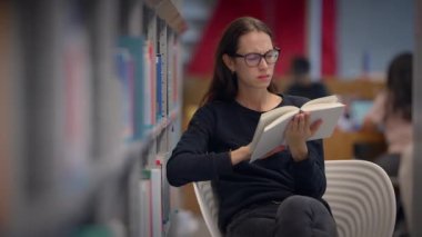 Kendini işine adamış bir kadın, çağdaş ve şık bir kütüphane ortamında kitap okumaya dalmıştır.