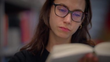 Genç bir kadın sessizce oturur ve bir kütüphane ortamında büyüleyici bir kitaba dalmış bir şekilde dikkatle okur.