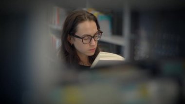 Genç bir kadın, raflarla çevrili çağdaş bir kütüphane ortamında kitap okumaya dalmış.