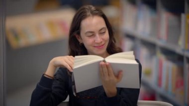 Neşeli genç bir kadın, rahat bir kütüphane ortamında rahatlarken büyüleyici bir kitaptan zevk alıyor.