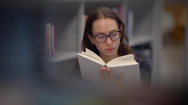 Kendini işine adamış ve odaklanmış bir kadın dingin bir kütüphane ortamında büyüleyici bir kitaba dalmış.