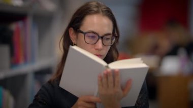 Gözlük takan bir kadın, uzun ve kütüphane raflarıyla çevrili bir kitabı okumaya dalmıştır.