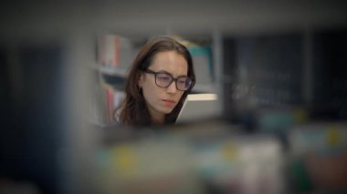 Gözlüklü bir kadın, uzun kitaplarla çevrili bir kütüphanede kitap okumaya dalmıştır.