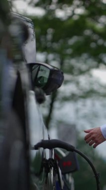 Park halindeki elektrikli bir araç, elektrik şarj aletine bağlanıyor. Hepsi de canlı, yemyeşil bir alanla çevrili.