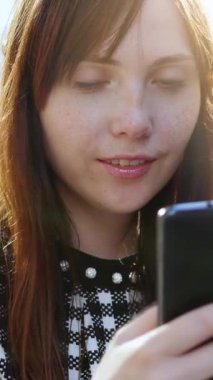 Genç bir kadın açık havada akıllı telefonunu kullanarak gülümsüyor. Dünyayla olan sevgi dolu bir anı yakalıyor.