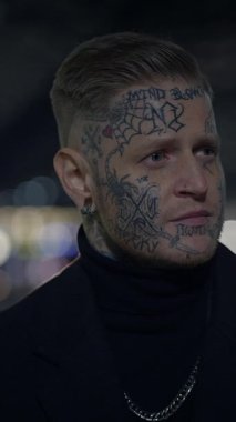 Odaklanmış bir adam karmaşık ve detaylı yüz dövmeleri sergiliyor, canlı bir şehrin arka planında geçiyor.