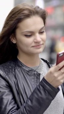 Genç bir kadın akıllı telefonunu enerji dolu, hareketli bir şehir ortamında kullanırken mutlu bir şekilde gülümsüyor.