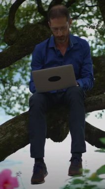 Dizüstü bilgisayarına derinden odaklanmış bir adam, bir ağaç dalına ustaca tünemiş, doğayı teknolojiyle mükemmel bir şekilde harmanlıyor.