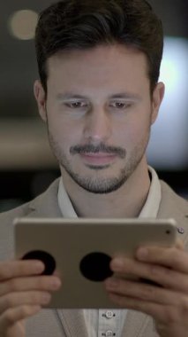 Terzi kıyafeti giymiş bir adam, şık, iyi ışıklandırılmış ve modern bir ortamda bir tablete odaklanır.