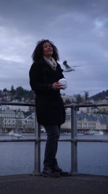 Bir kadın, bulutlu gökyüzünün güzel bir zeminiyle çerçevelenmiş belli bir nesneyi tutarken suyun kenarında duruyor.