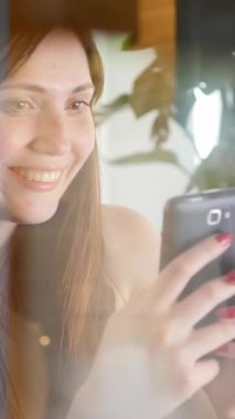 Neşeli bir kadın, sıcak ve davetkar doğal ışıkla aydınlatılırken, akıllı telefonuyla mutlu bir şekilde etkileşim içindedir.