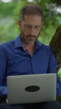 Bir adam laptopuna dalmış, açık havada rahatça otururken, tamamen yemyeşil ile çevrili.