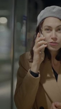 Moda beresi takan şık bir genç kadın gece canlı bir şehir ortamında cep telefonuyla konuşuyor.