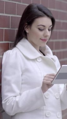 Şık beyaz önlüklü şık bir kadın, akıllı telefonuyla tuğla bir duvarın önünde dikilirken meşgul oluyor.