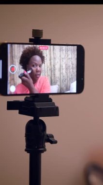 Parlak pembe gömlekli bir kadın akıllı telefon cihazını kullanarak bir video kaydederken saçlarını nazikçe düzeltiyor.