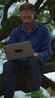 Bir adam ağaçta rahatça oturur, dizüstü bilgisayarıyla çalışır, doğa ve verimliliği kusursuz bir şekilde harmanlar.