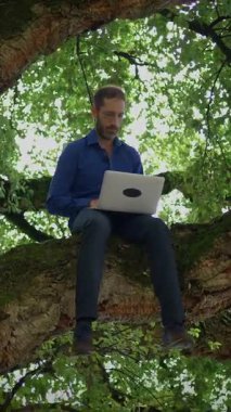 Bir insan bir dizüstü bilgisayarla çalışırken ağaçta rahatça oturuyor ve doğayı modern teknolojiyle harmanlıyor.