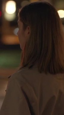 Bir kadın, yumuşak ışıkların büyülü bir atmosfer yarattığı bir şehirde, akşam gökyüzünün altında düşünceli bir şekilde duruyor.