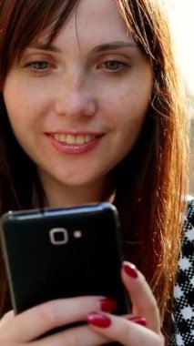 Neşeli bir kadın, akıllı telefonuyla hoş bir şekilde meşgul, açık havada güneşin tadını çıkarıyor.