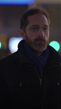 Şık bir ceket ve eşarp giyen düşünceli bir adam gece boyunca canlı ve canlı bir şehir ortamında duruyor.